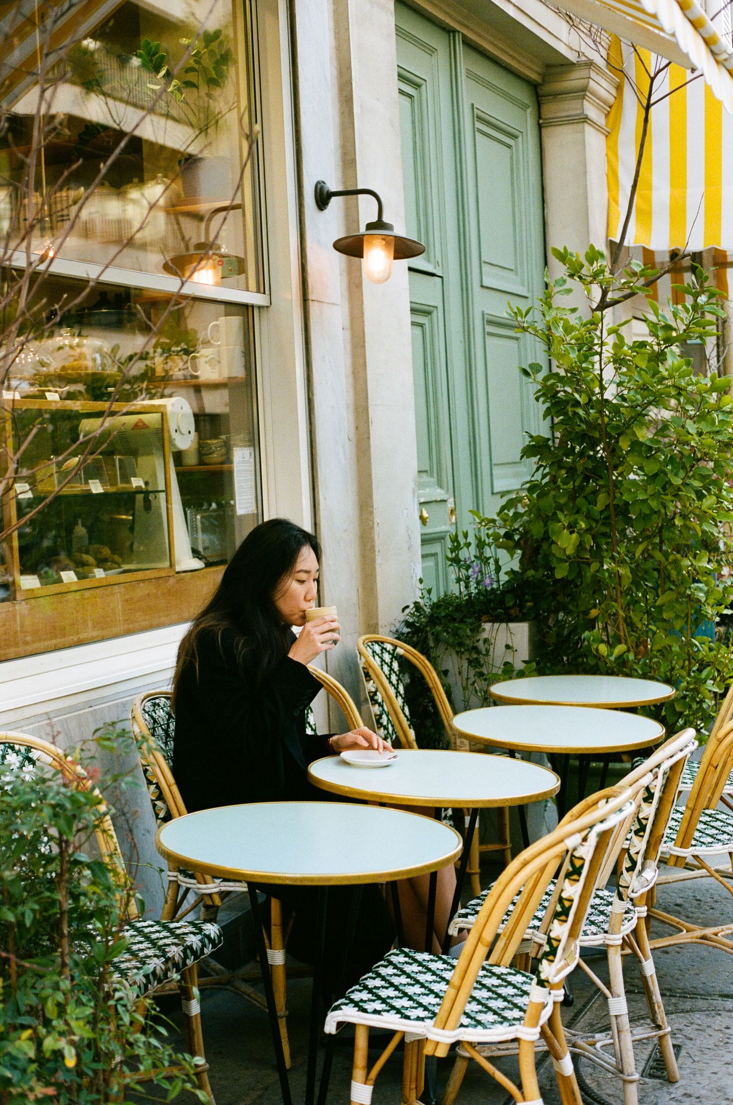 A cafe scene with patterned chairs, round marble tables and green potted plants against a mint green exterior.