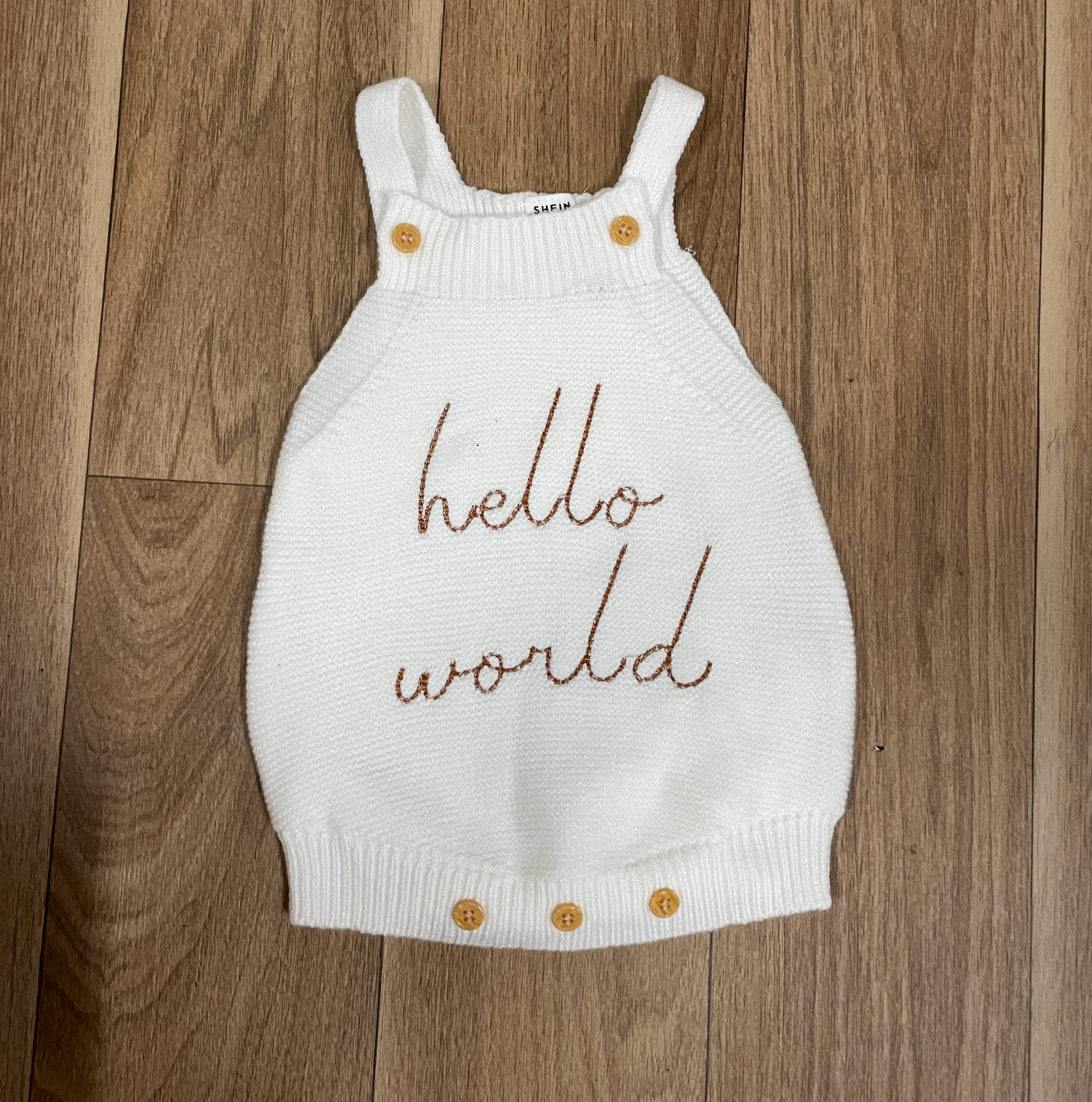 White knit baby top with 'hello world' text embroidered in brown thread laying on wooden floor.