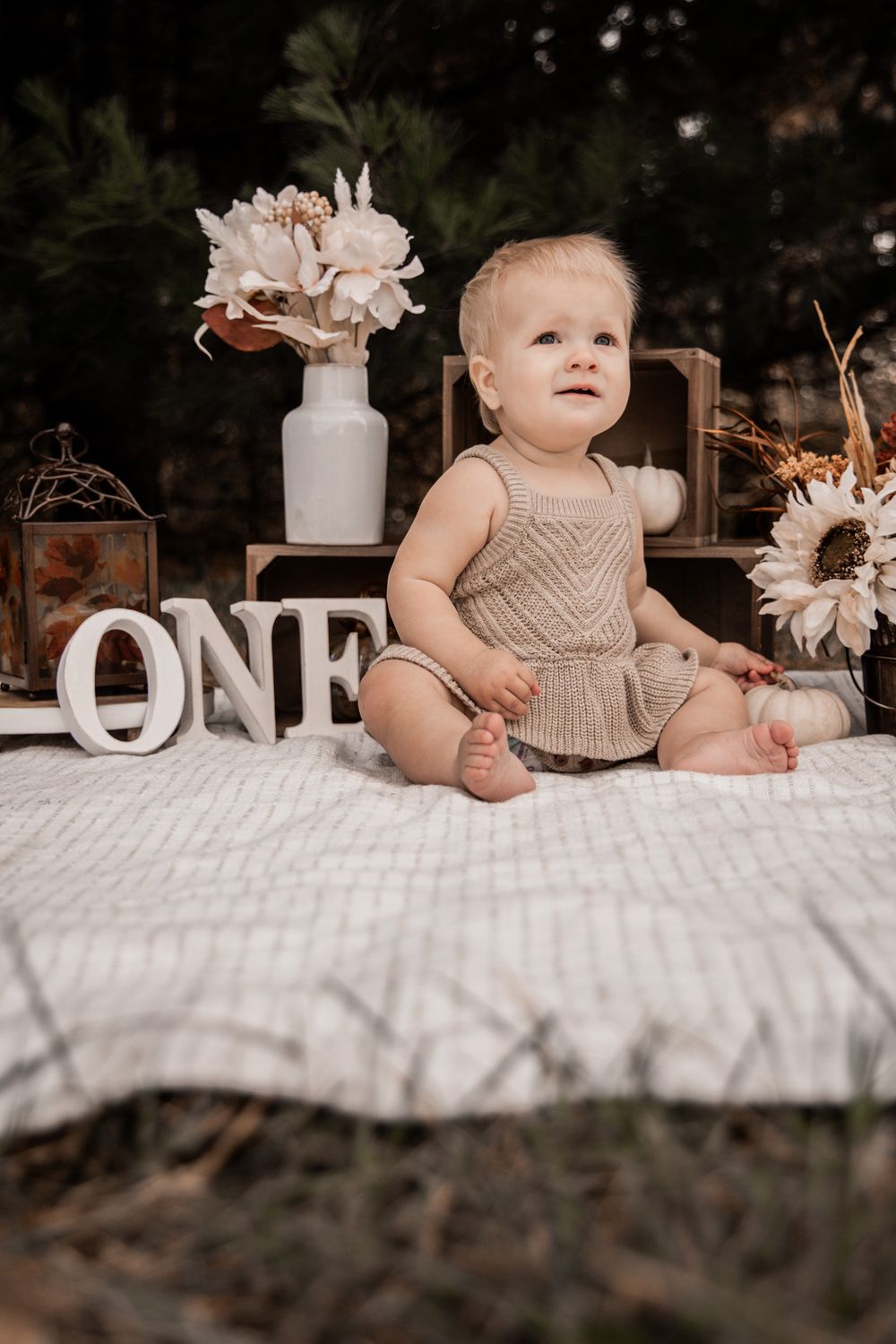 A first birthday photo setup with white decor, floral arrangements, and a baby in a beige romper sitting on a blanket.