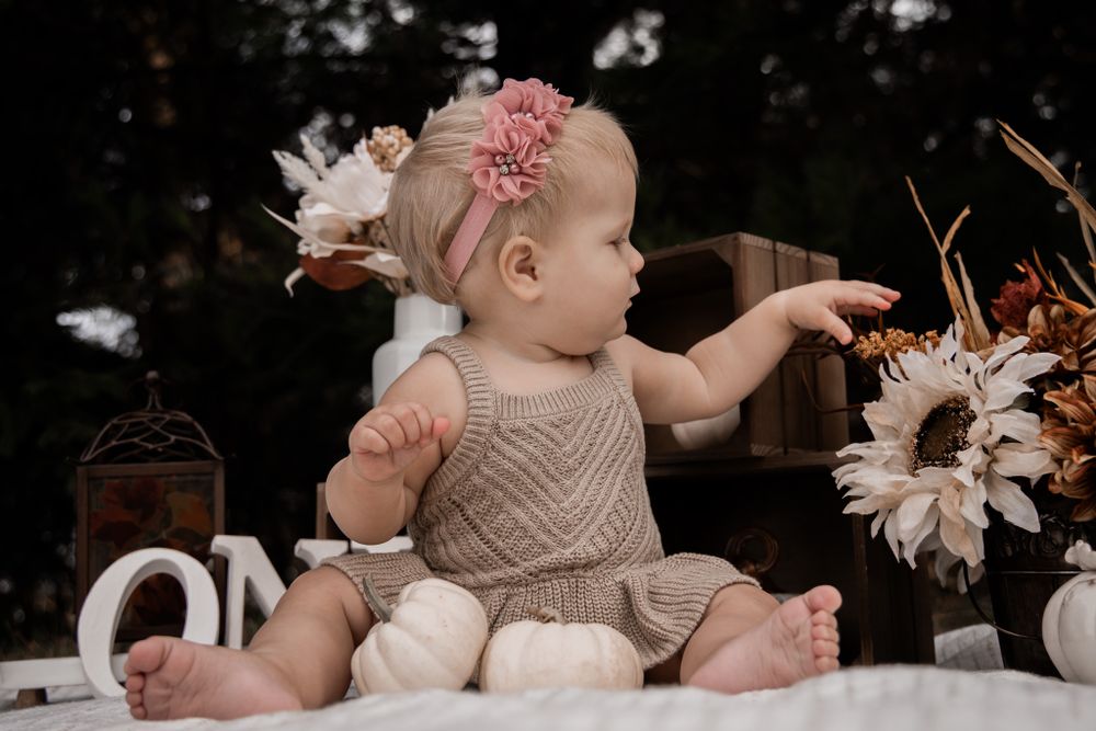 A baby in beige clothing reaches for autumn-themed decorations during their first birthday photoshoot.
