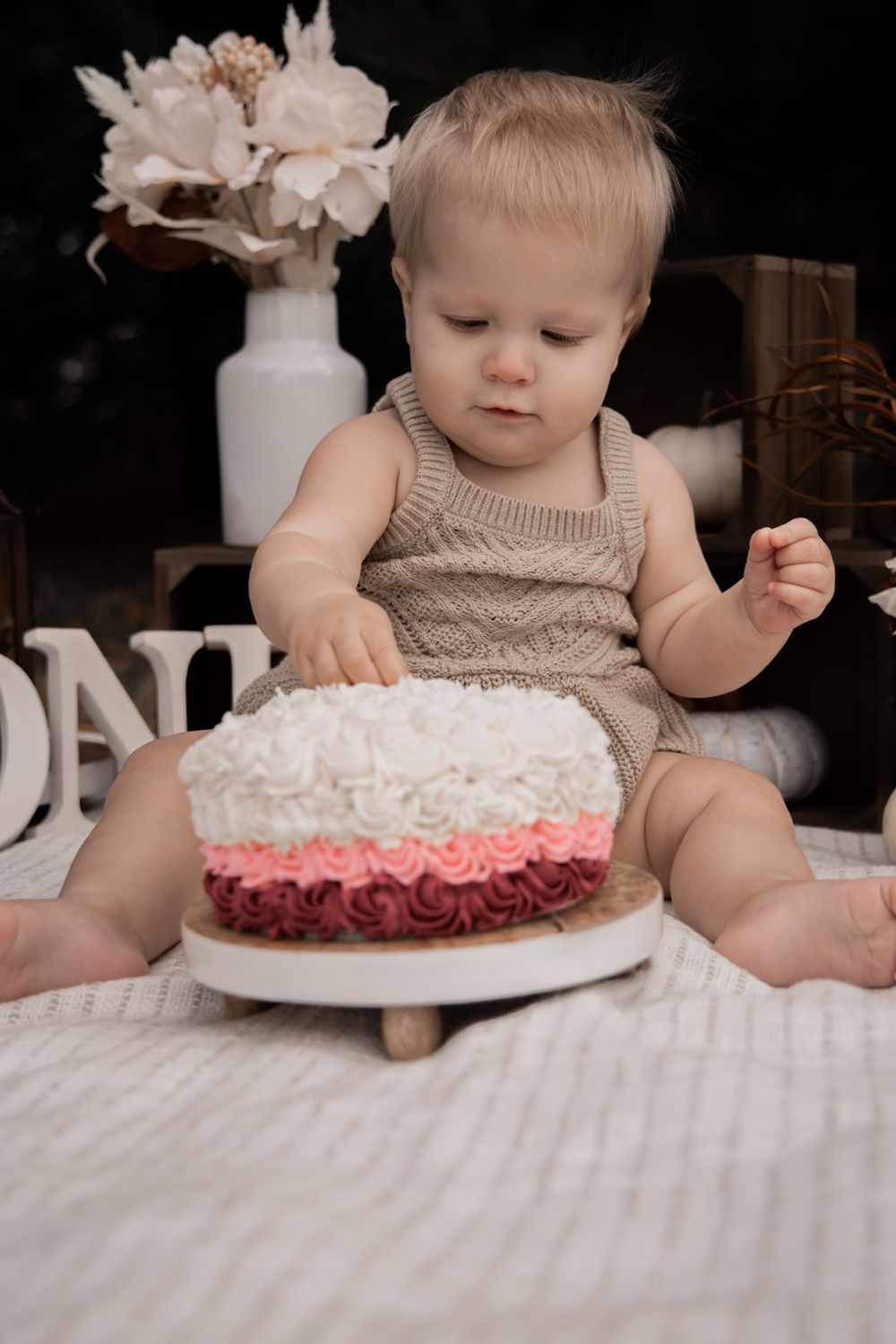 Baby in beige clothing examines a white and pink frosted birthday cake during first birthday celebration.