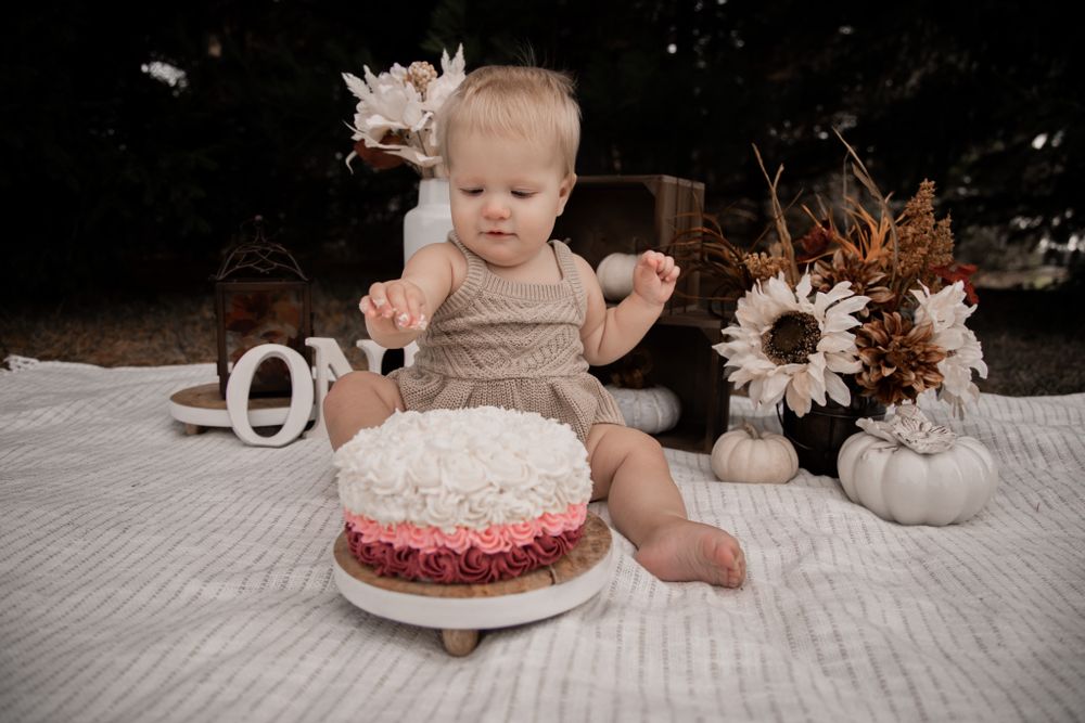 First birthday celebration with baby in beige clothing sitting near a decorated cake on white blanket.