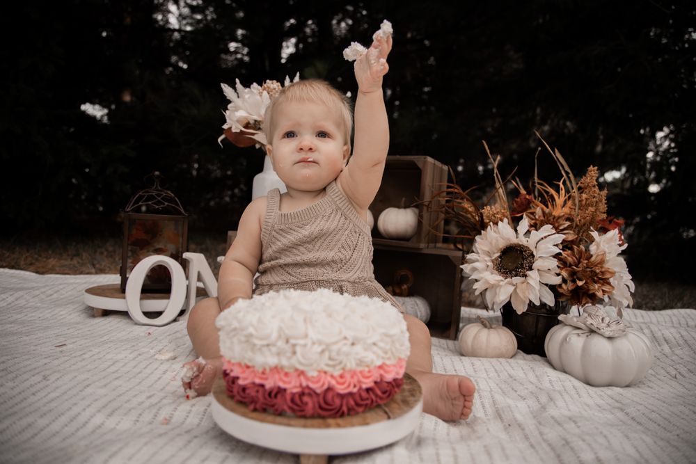 A baby in a tan romper reaches up while sitting next to a pink and white cake and decorative pumpkins on a blanket.