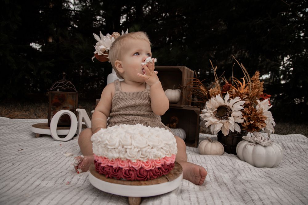 A baby in beige clothing enjoying their first birthday cake with white and red frosting decorations.