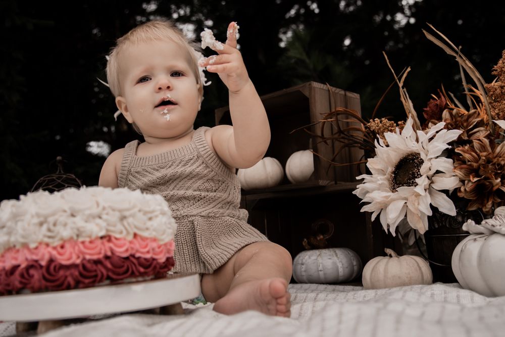 Baby with raised hands sits on white blanket beside ombre cake and fall decor.