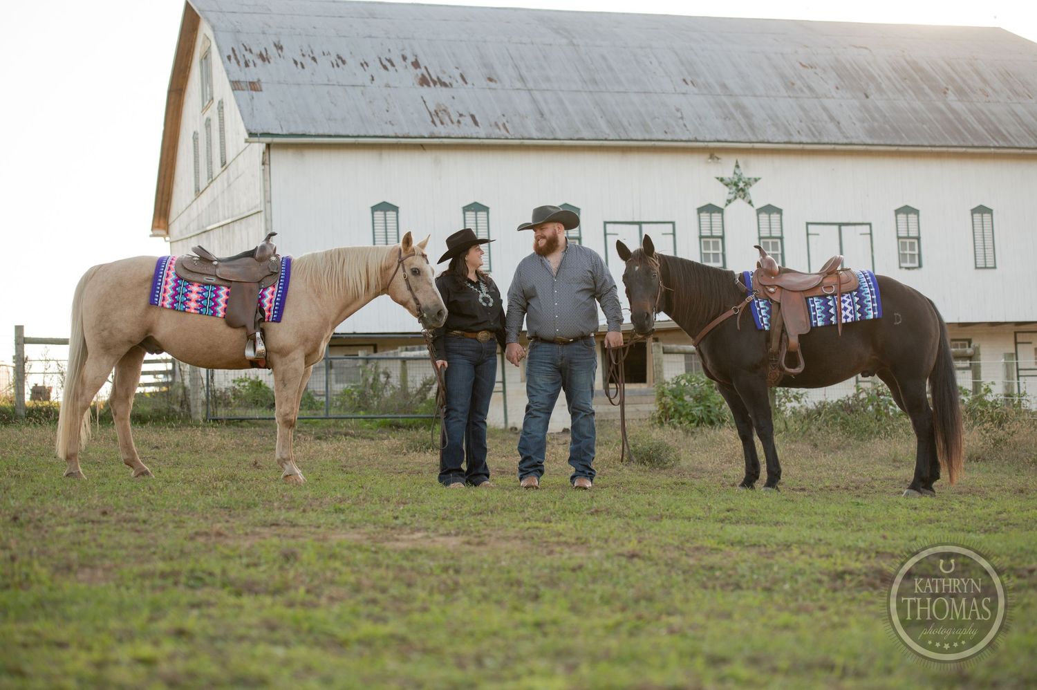 Western-dressed couple stands between two saddled horses by white barn.