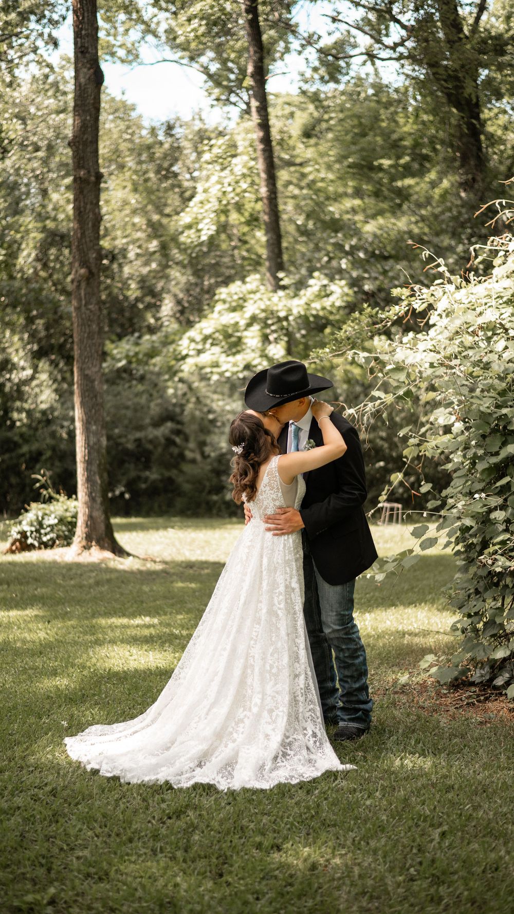 A couple in a white lace wedding dress and western attire share an embrace in a sunlit forest setting.