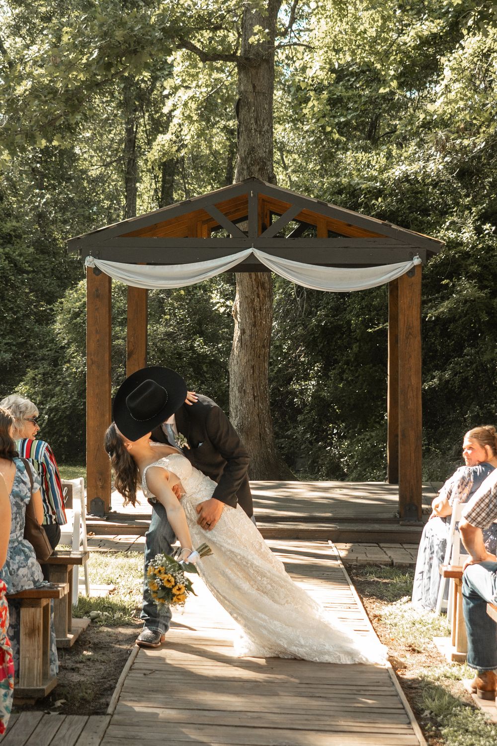 A romantic dip kiss under a wooden wedding arbor draped with white fabric in a sunlit forest setting.