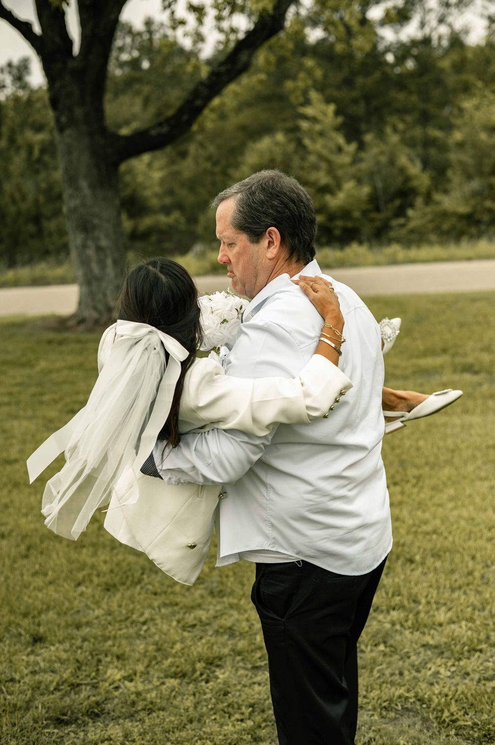 A bride and older man share an emotional embrace outdoors near a large tree during a wedding ceremony.