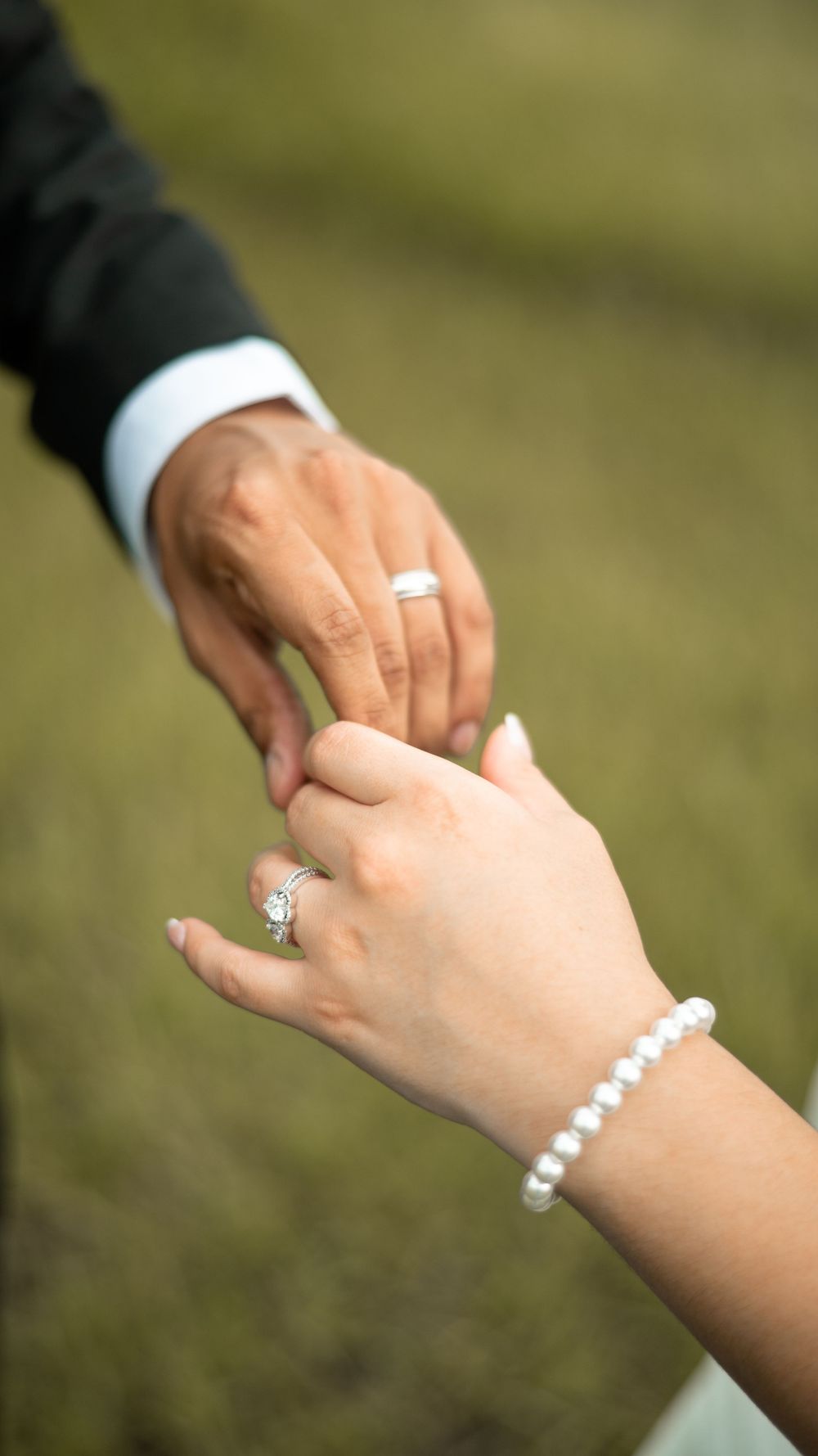 Two hands wearing wedding rings and a pearl bracelet touch tenderly against a blurred green outdoor background.