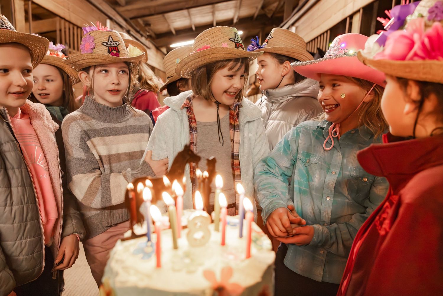 A group in cowboy hats gather around a glowing birthday cake with lit candles in a rustic indoor setting.