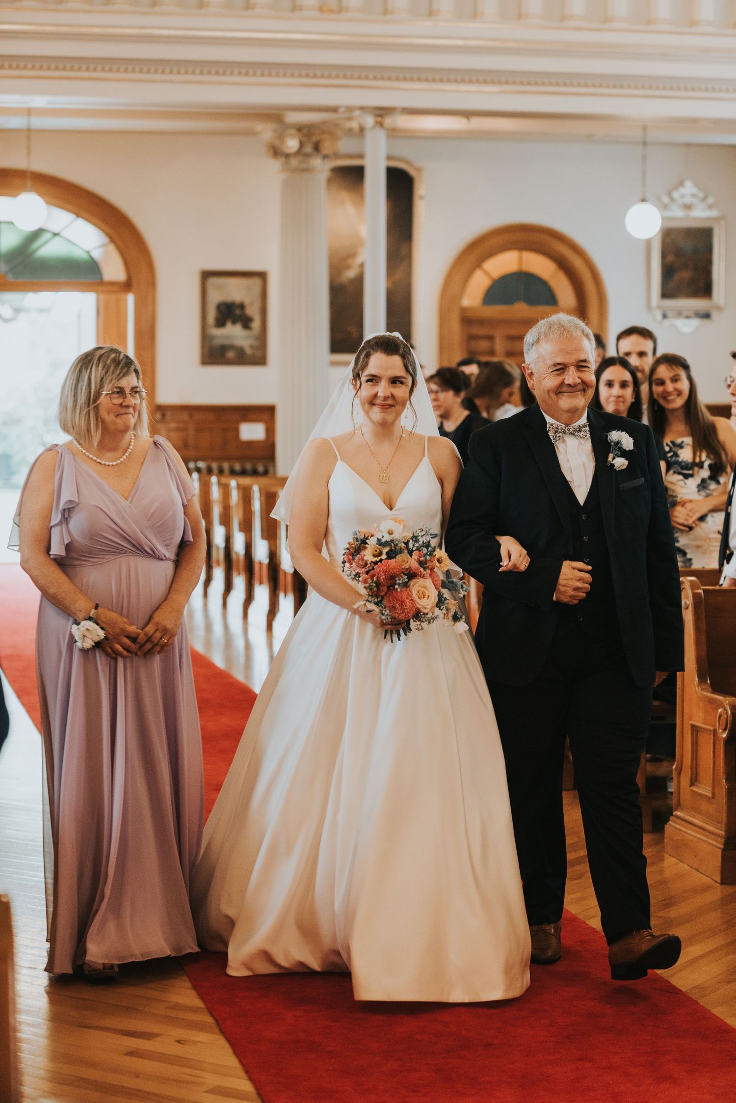 A bride in a classic white gown walks down the aisle on a red carpet escorted by a companion during a traditional ceremony.