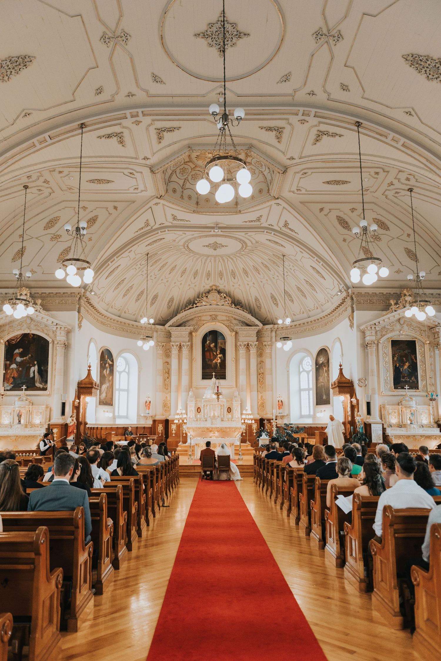 Interior view of an ornate Catholic church with vaulted ceiling, elegant chandeliers and wooden pews along a red aisle runner.