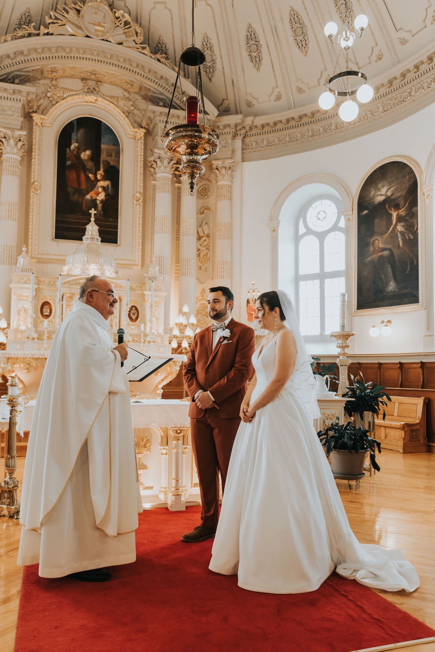 A traditional wedding ceremony taking place in an ornate cathedral with white and gold decor and religious paintings.