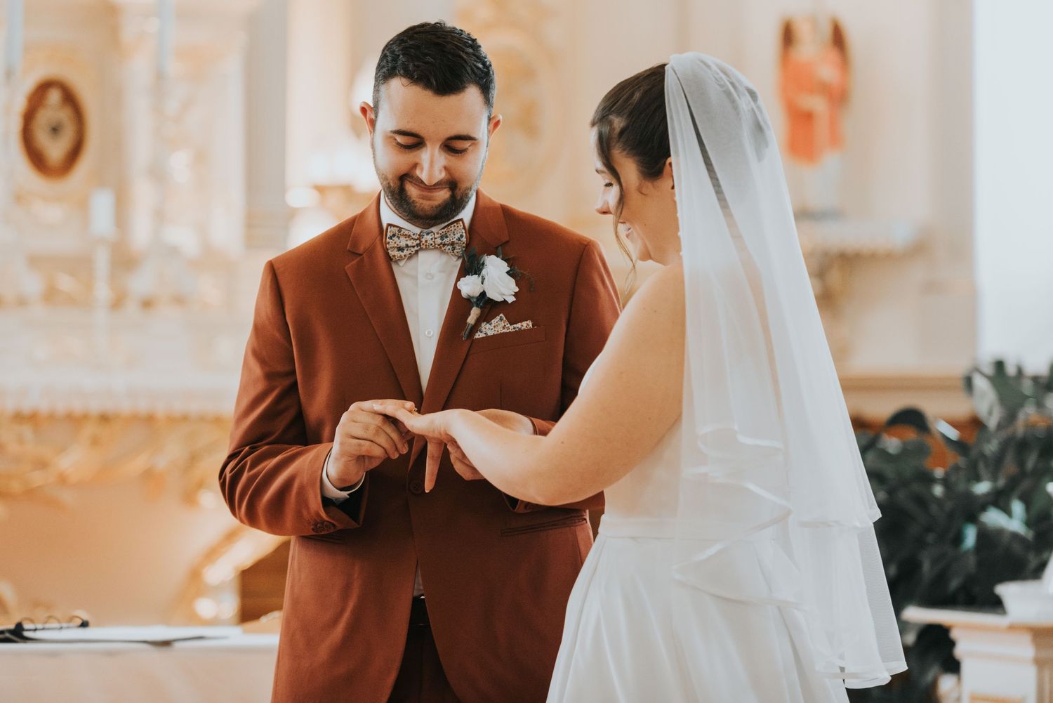 A couple exchanges wedding rings during their ceremony in an elegant church with ornate white decor.