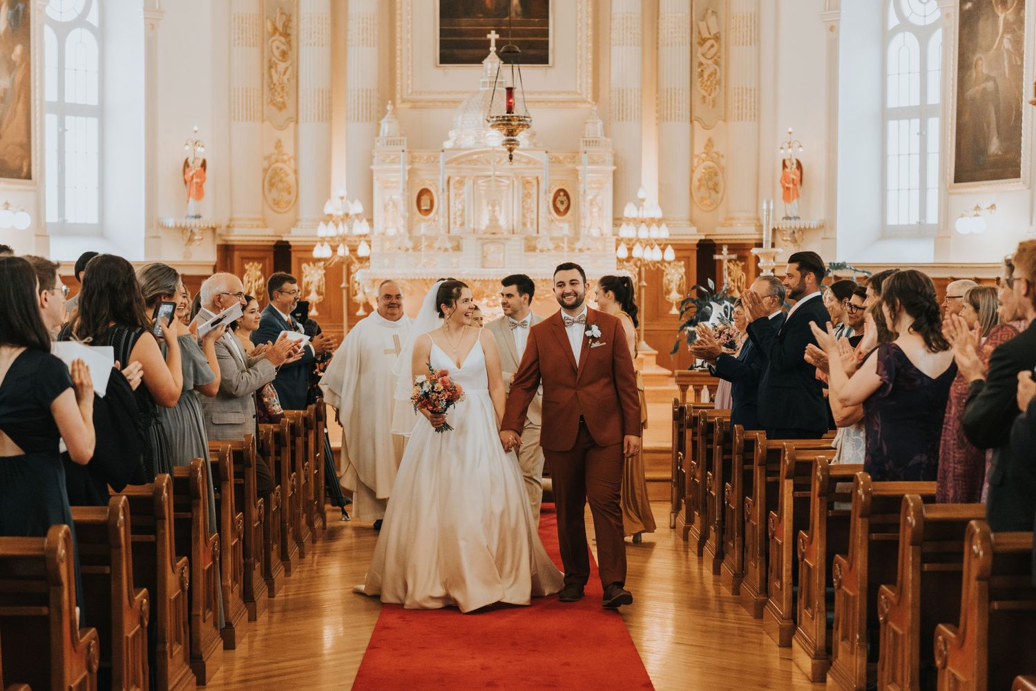 A couple walks down the red-carpeted church aisle while wedding guests applaud from wooden pews.