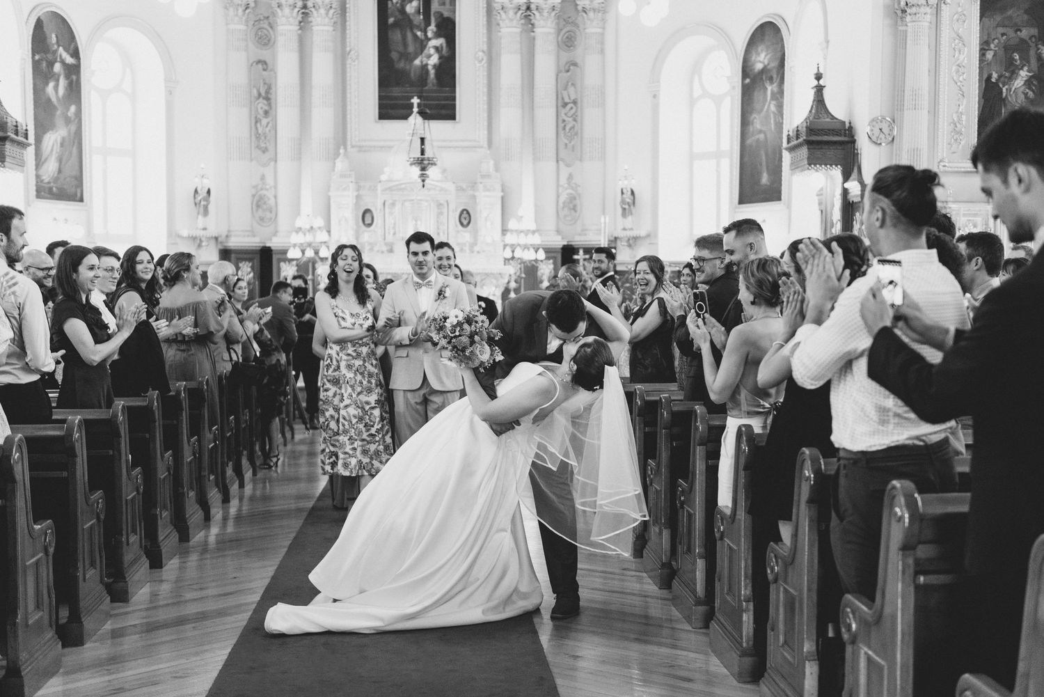 Elegant wedding celebration in a bright cathedral church with guests lined up along wooden pews in black and white.