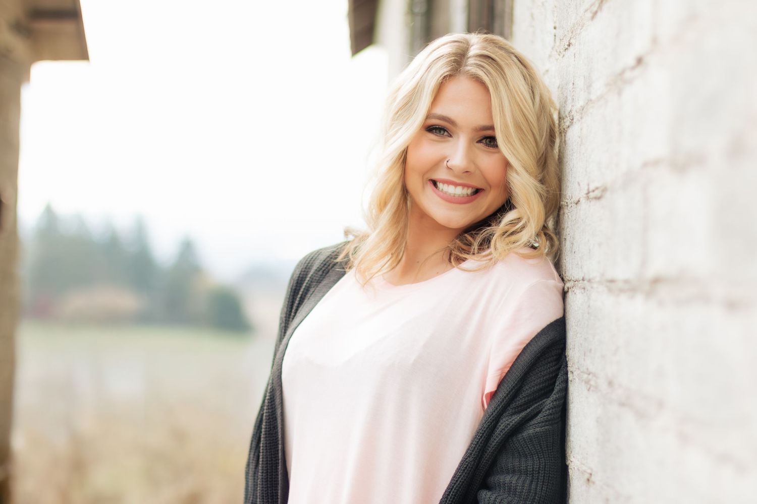 A happy woman with blonde hair leans against a white wall wearing a white shirt and dark cardigan outdoors.