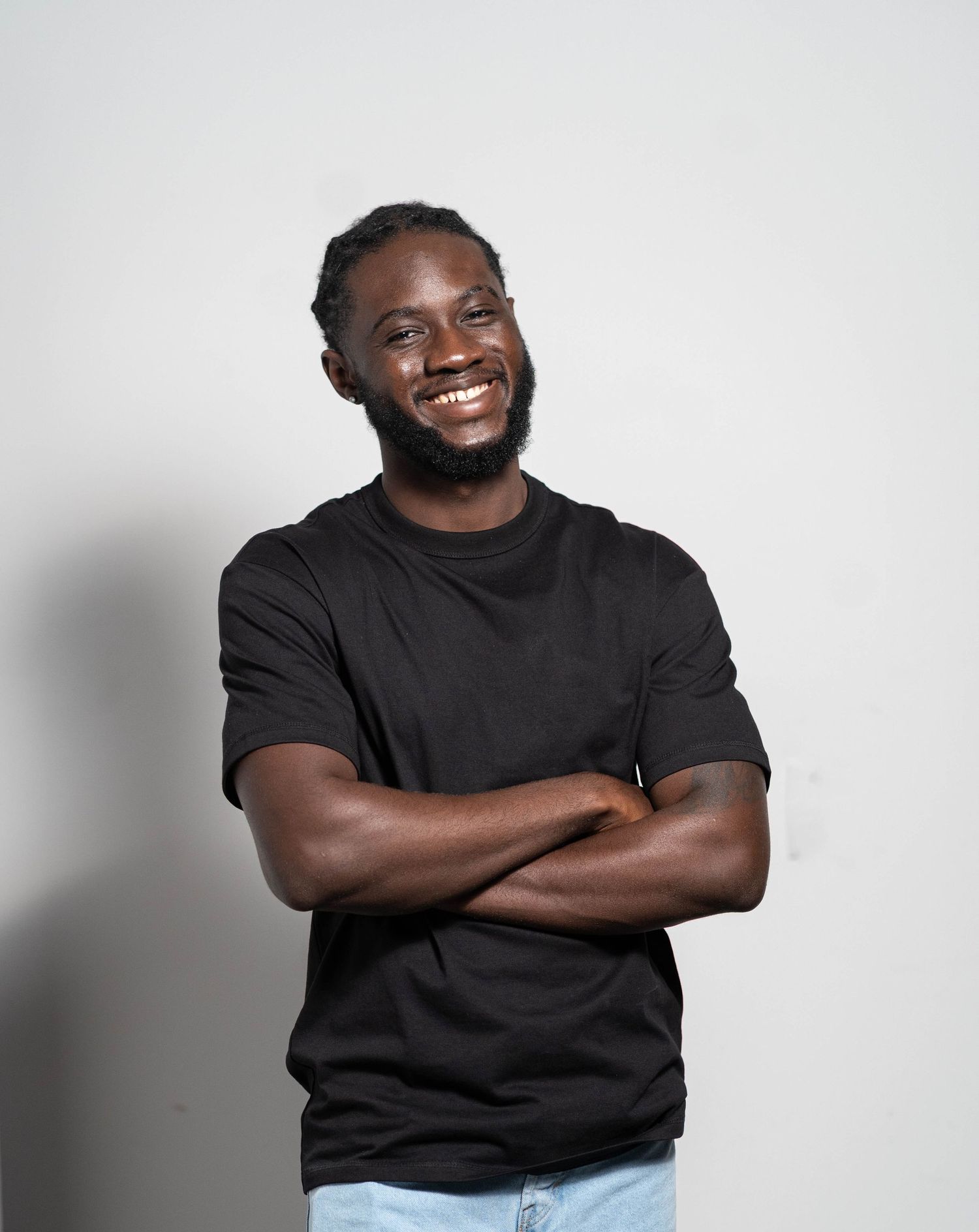 Person wearing a black t-shirt stands with arms crossed and a warm smile against a light gray background.