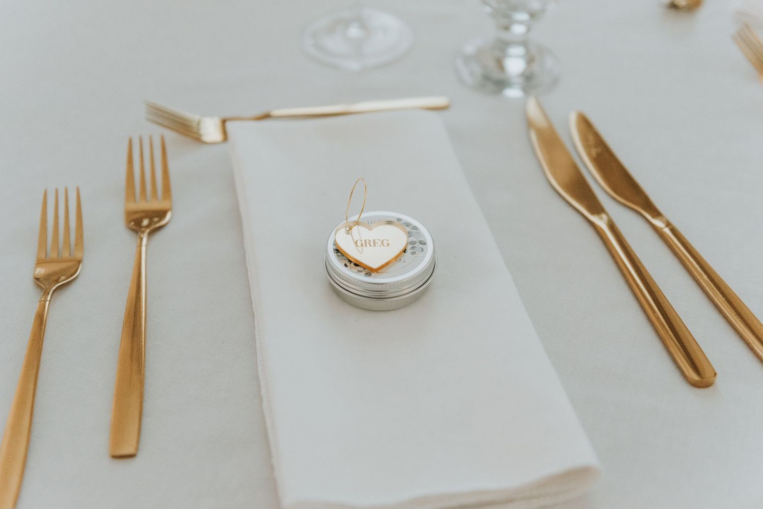 Elegant gold cutlery and a heart-shaped wedding favor arranged on a white linen napkin at a formal place setting.