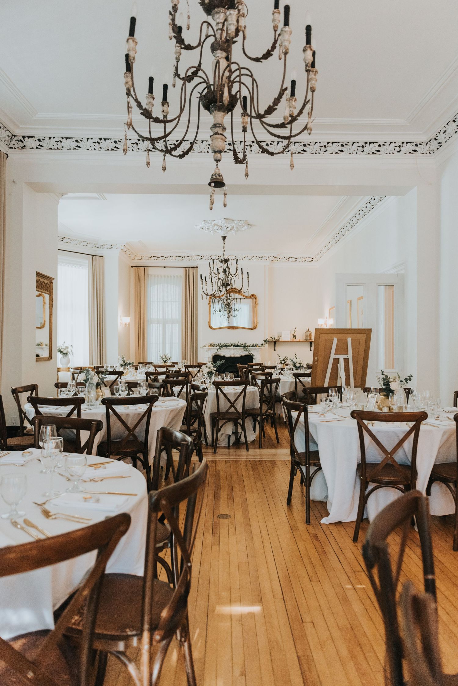 Elegant dining room with crystal chandeliers, white tablecloths, and dark wood cross-back chairs on hardwood floors.