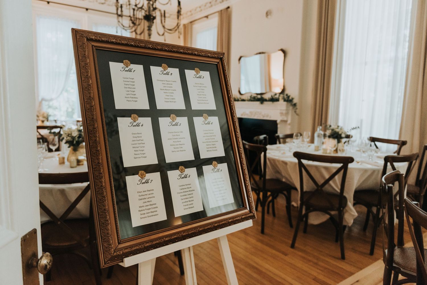 An elegant wedding seating chart displayed in an ornate gold frame on an easel in a classic dining room setting.