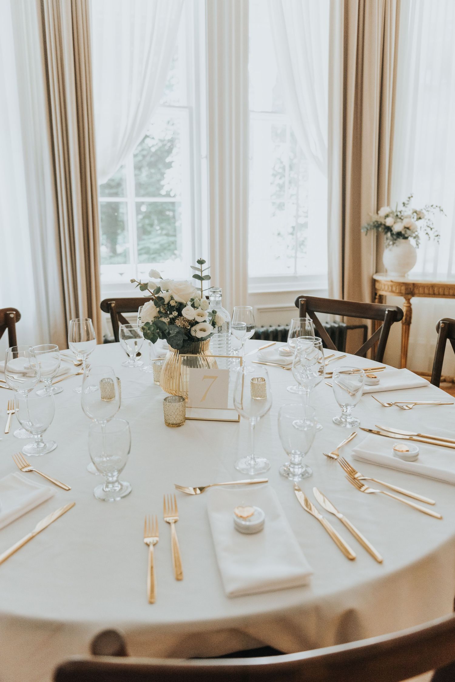An elegant wedding reception table setting with white linens, floral centerpieces and formal place settings in a bright room.