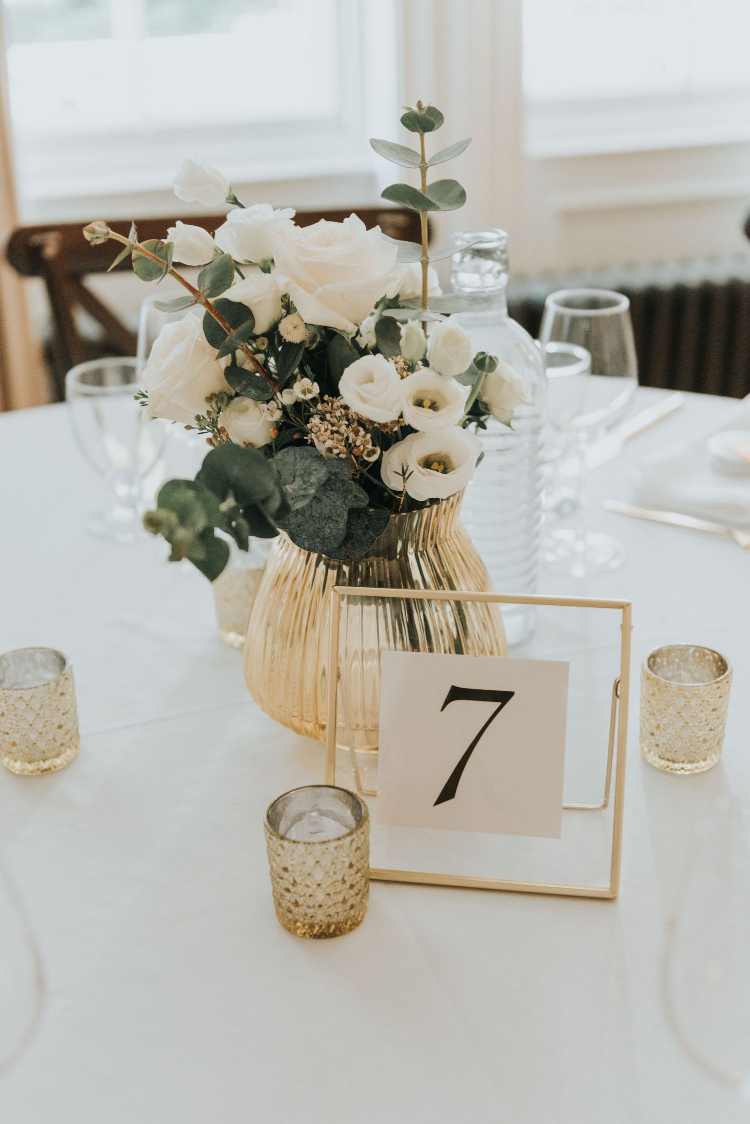 Modern white and gold table decor with table number 7, featuring white roses and eucalyptus in a gold vase.