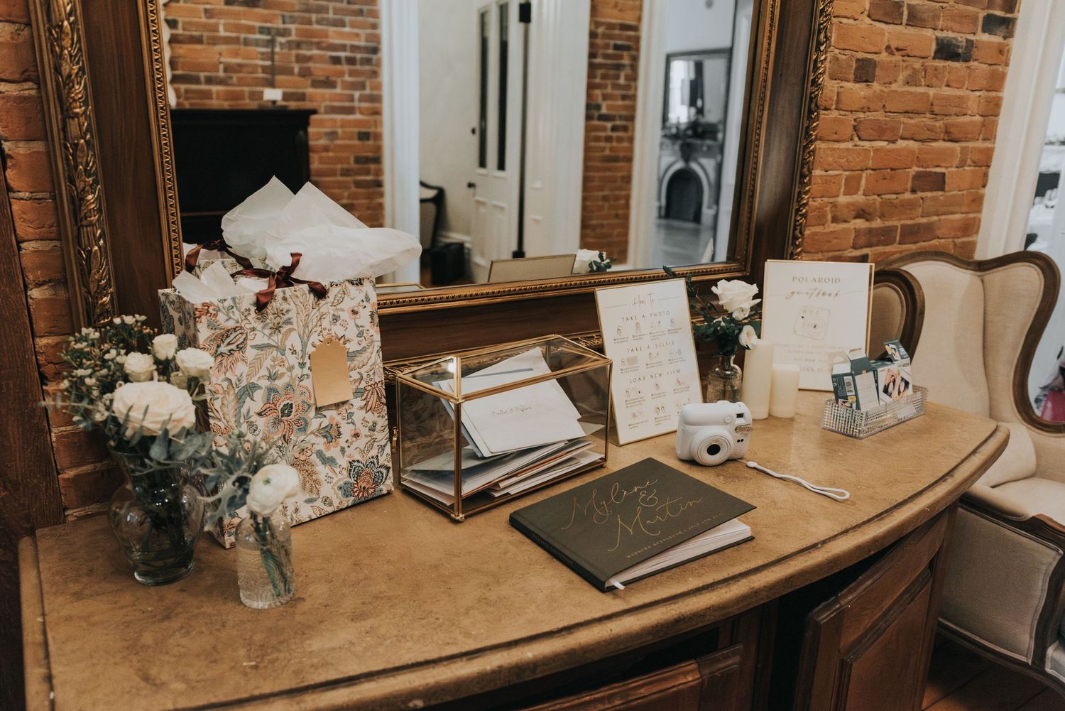 Vintage vanity desk with flowers, laptop, and wedding preparation items against exposed brick wall.