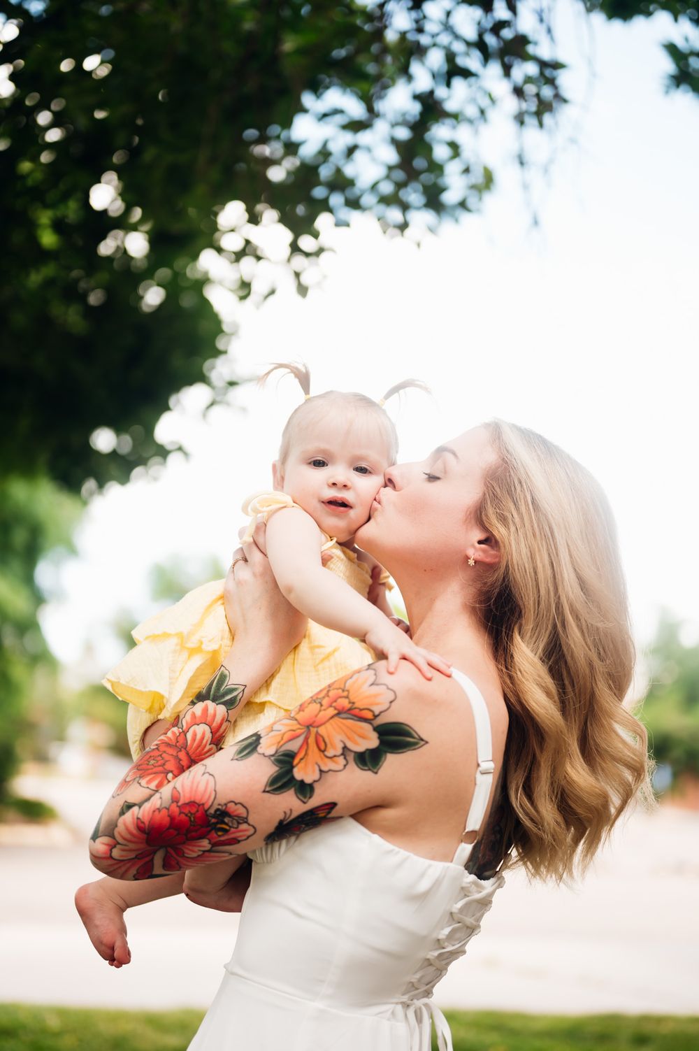 A joyful outdoor portrait shows a tender moment shared between parent and baby in sunny natural lighting.