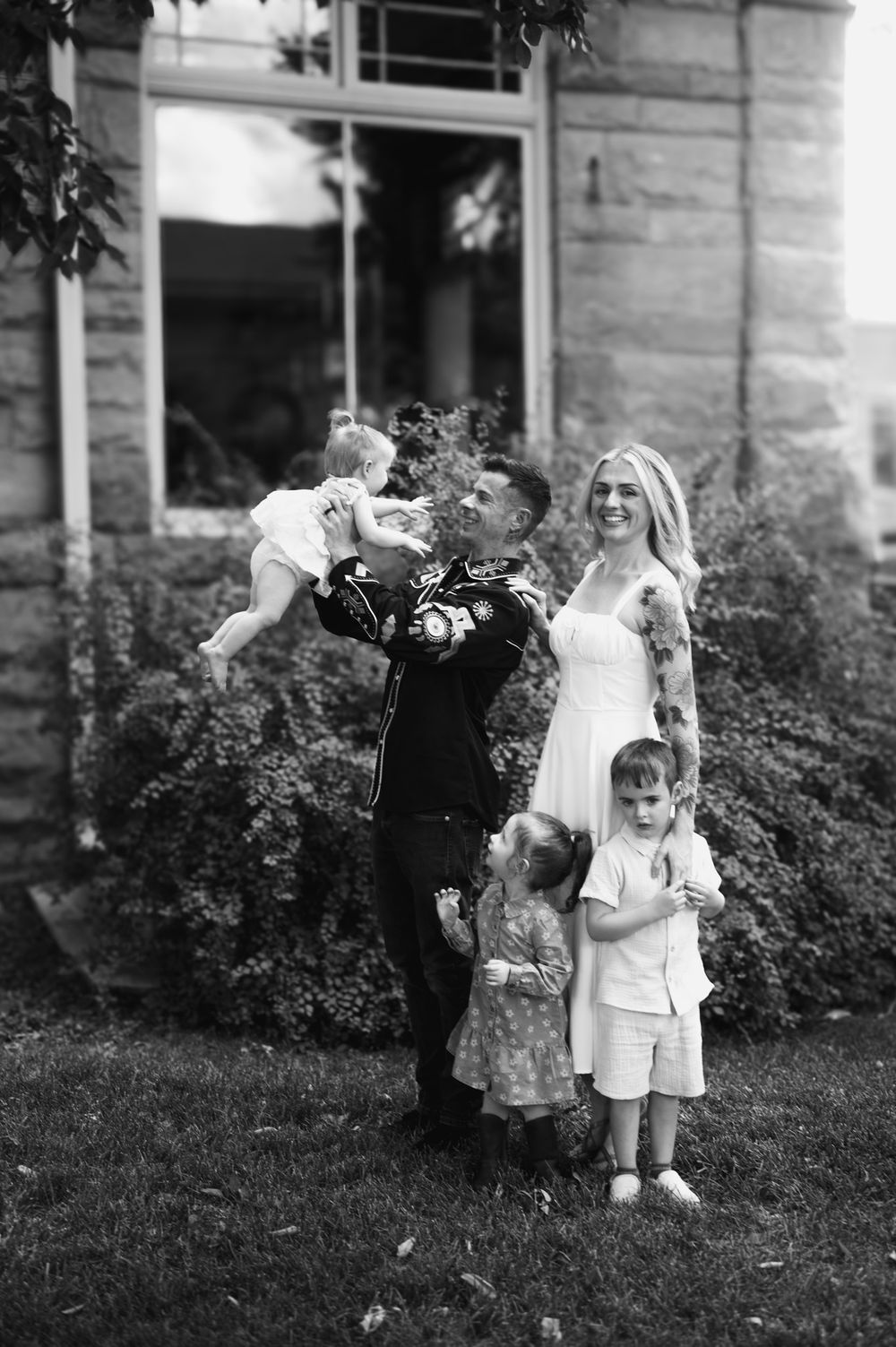 A black and white family portrait of a couple with children standing outdoors by a house with shrubs in the background.