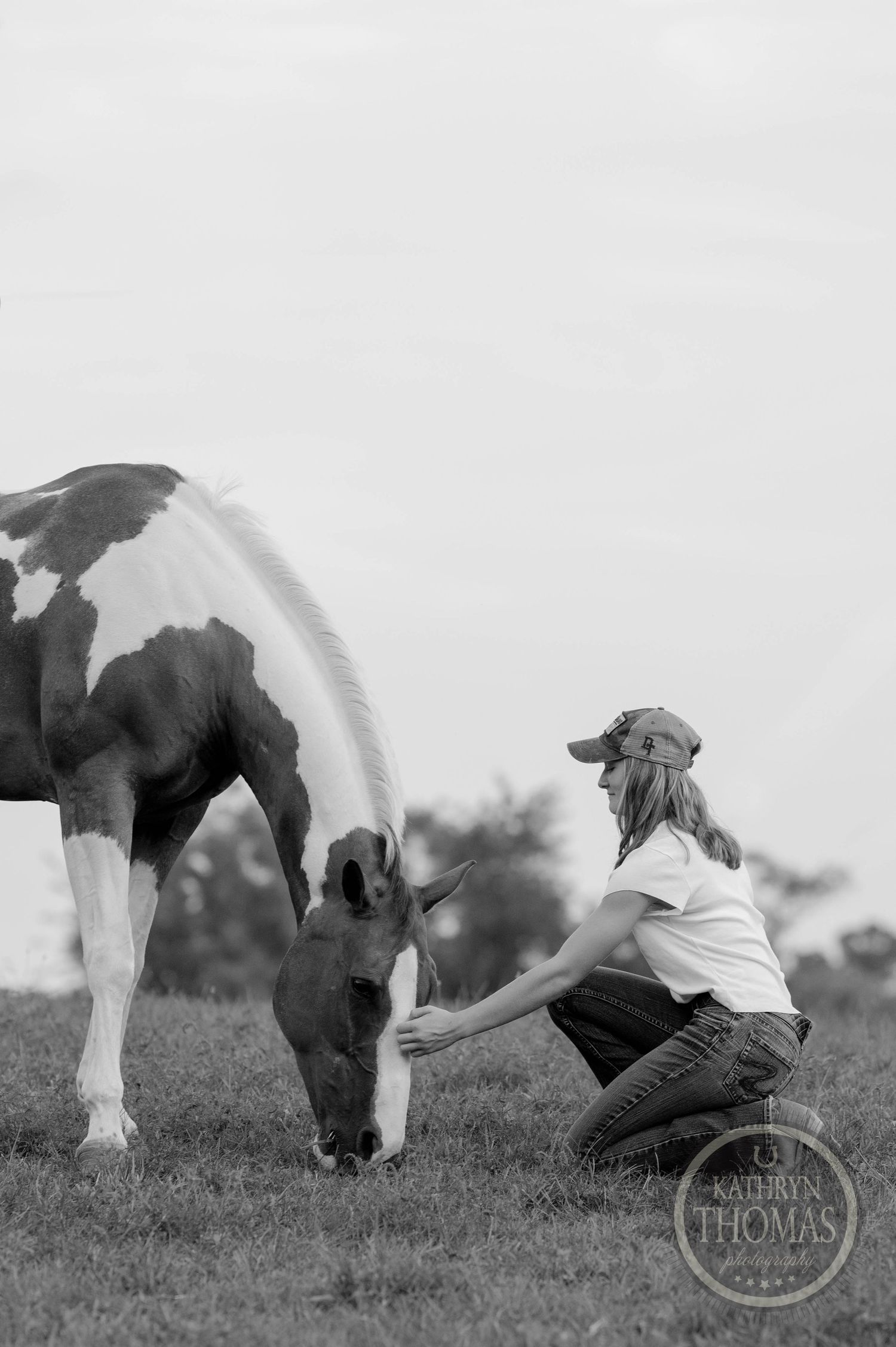 Black and white photo shows person petting paint horse while kneeling in grassy field.