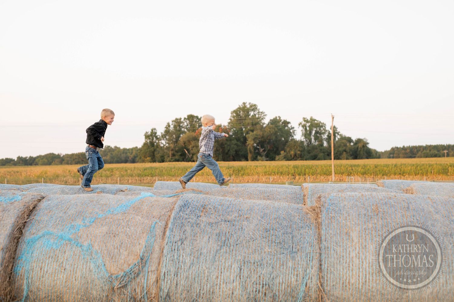 Two kids playing and jumping on rolled hay bales in a golden farm field.