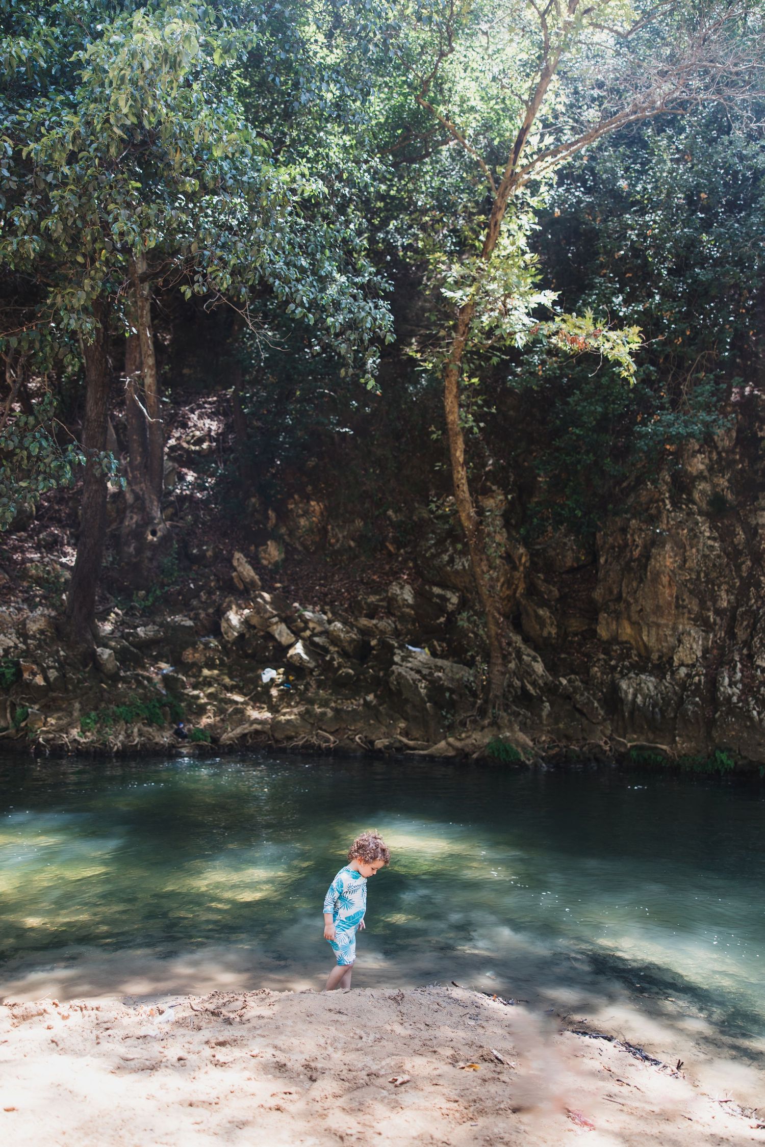 A child explores a serene swimming hole surrounded by rocky cliffs and lush trees.