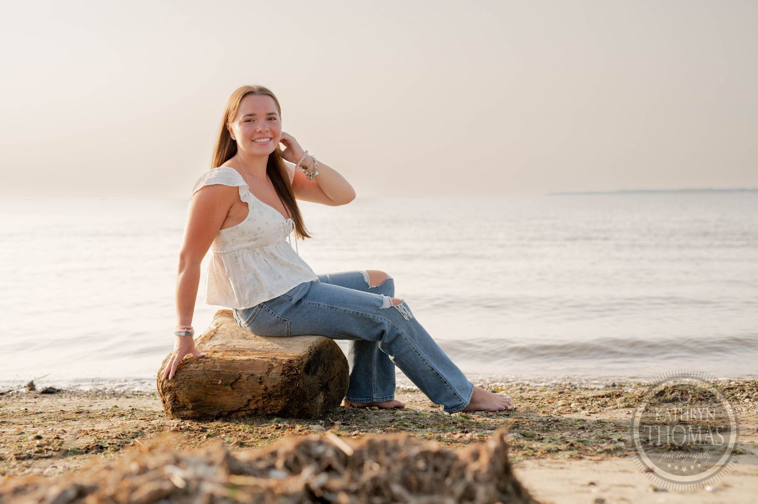 Person in white top and jeans sitting on a rock by the ocean during golden hour.