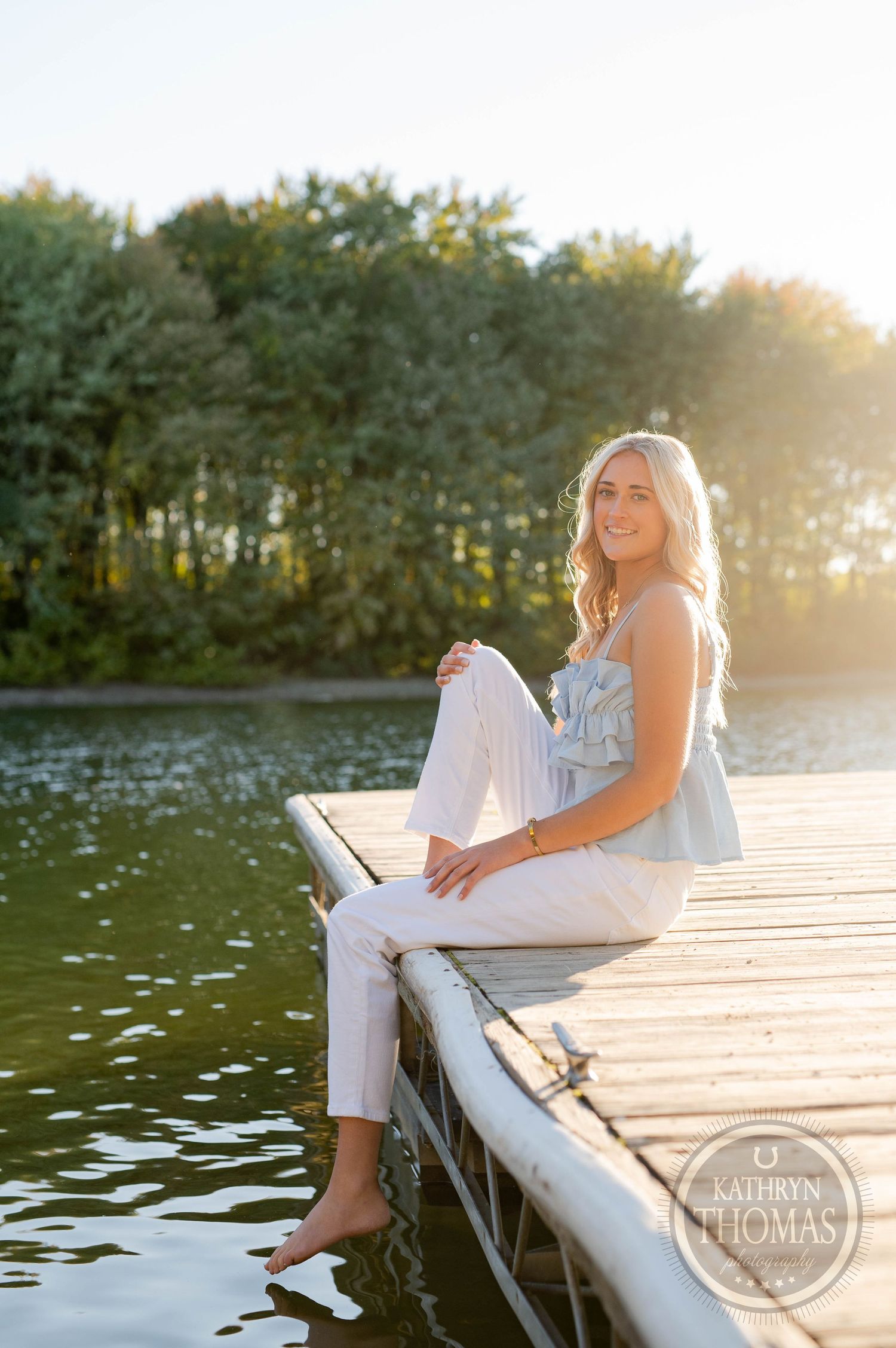 Summer portrait of someone in casual attire sitting on wooden dock by serene lake at sunset.