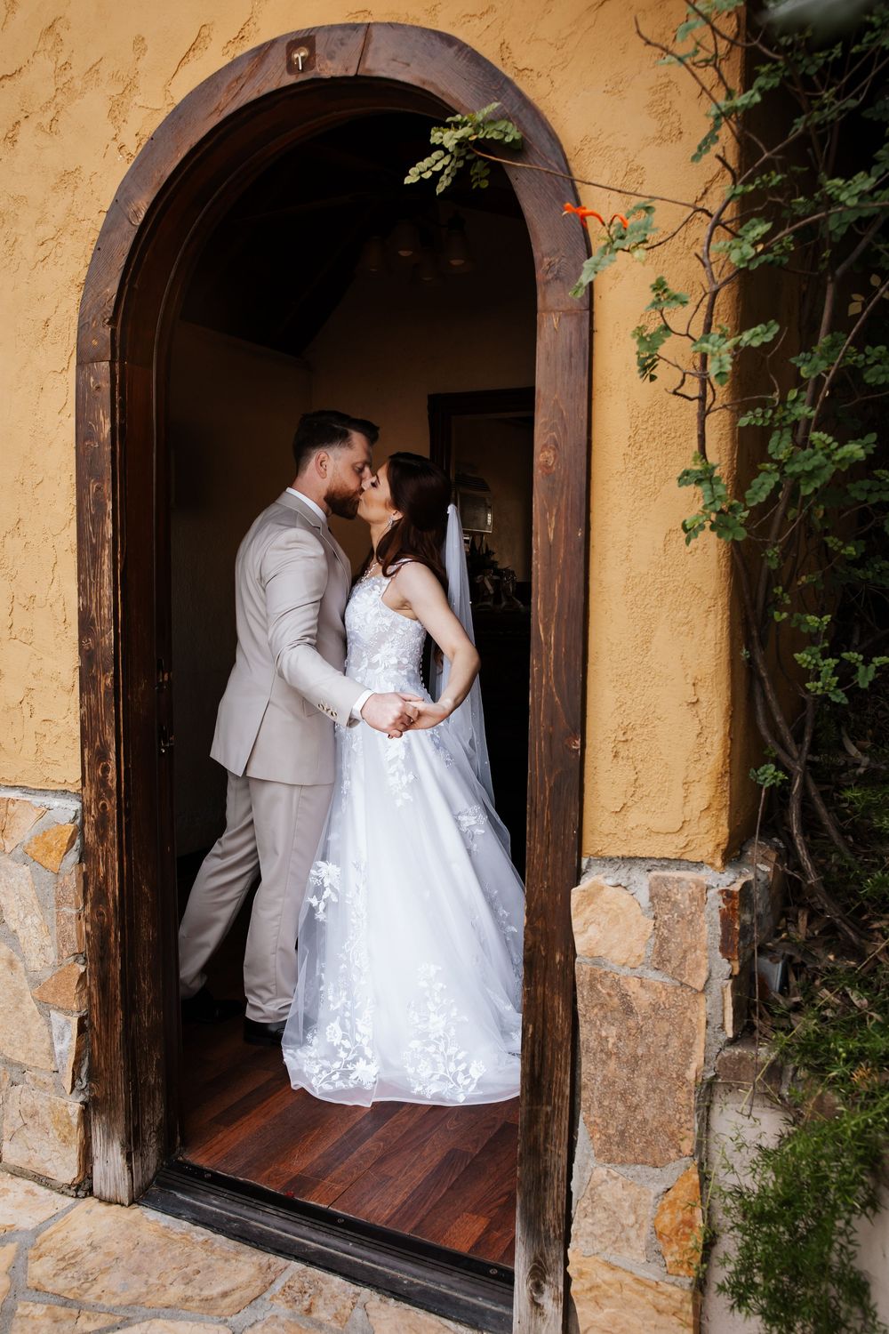 Bride and groom share a kiss framed by a charming arched wooden doorway of a chapel.