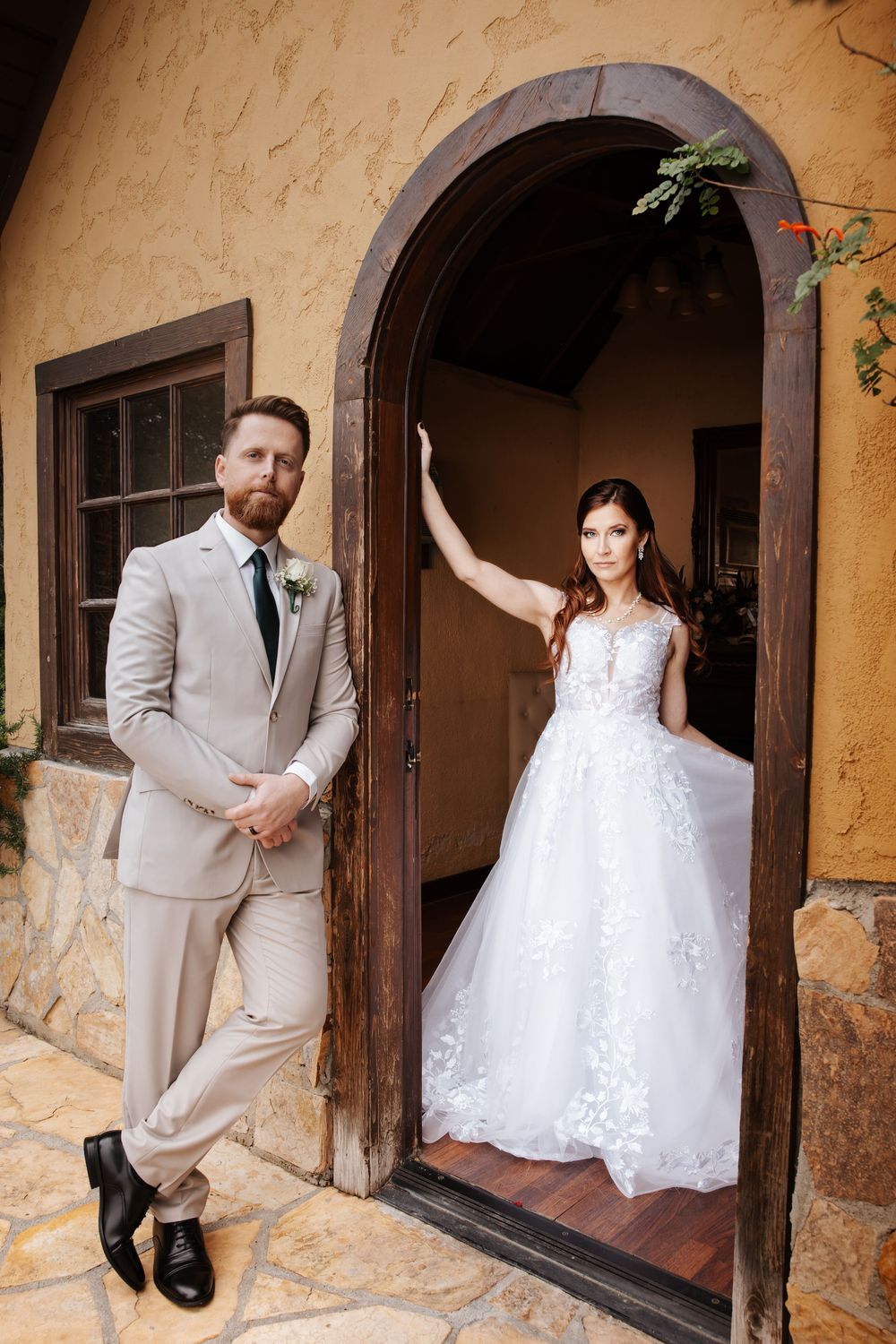 Groom in tan suit leans against chapel wall as bride poses elegantly in arched doorway.