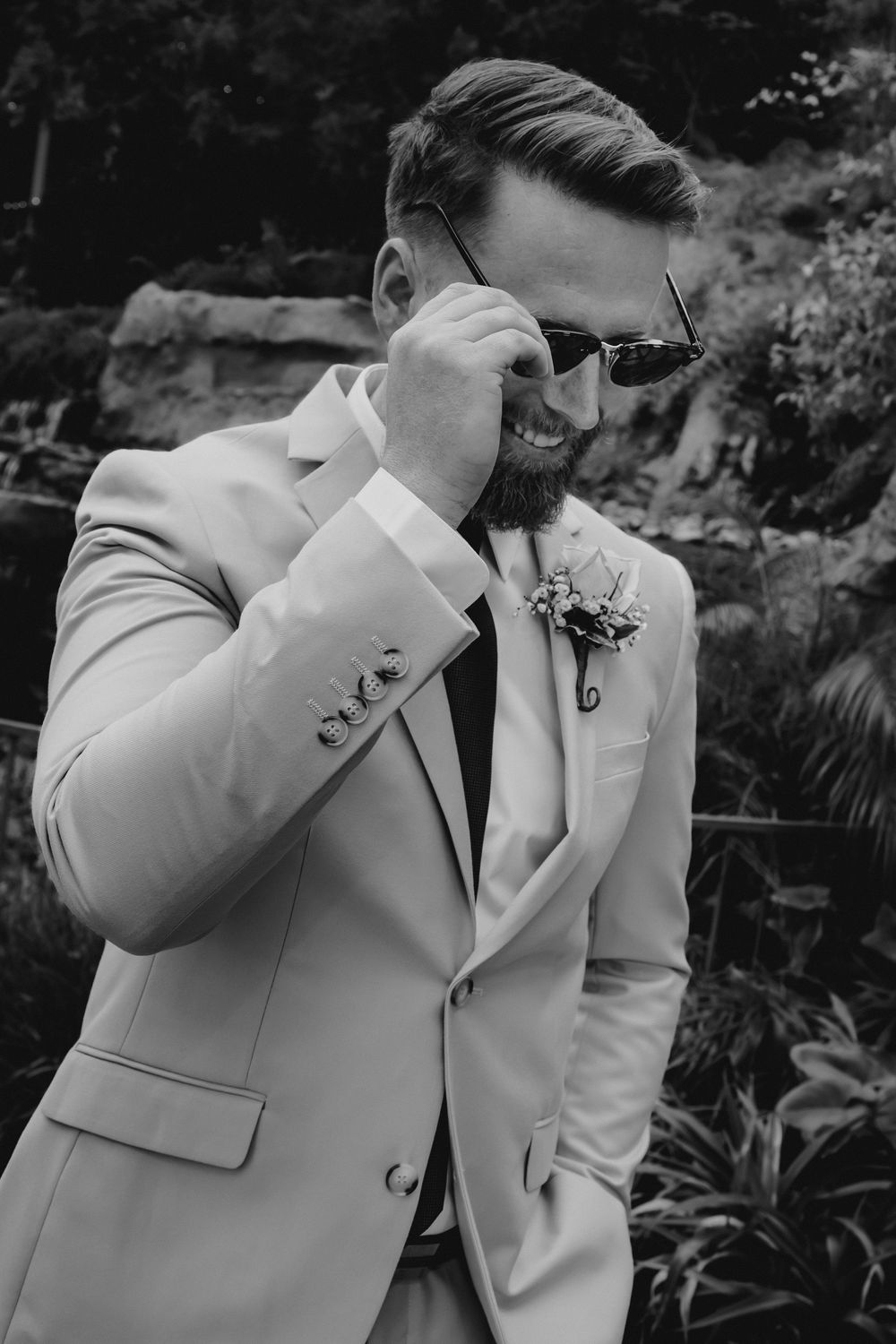 Black and white portrait of smiling groom in light suit adjusting sunglasses at wedding.