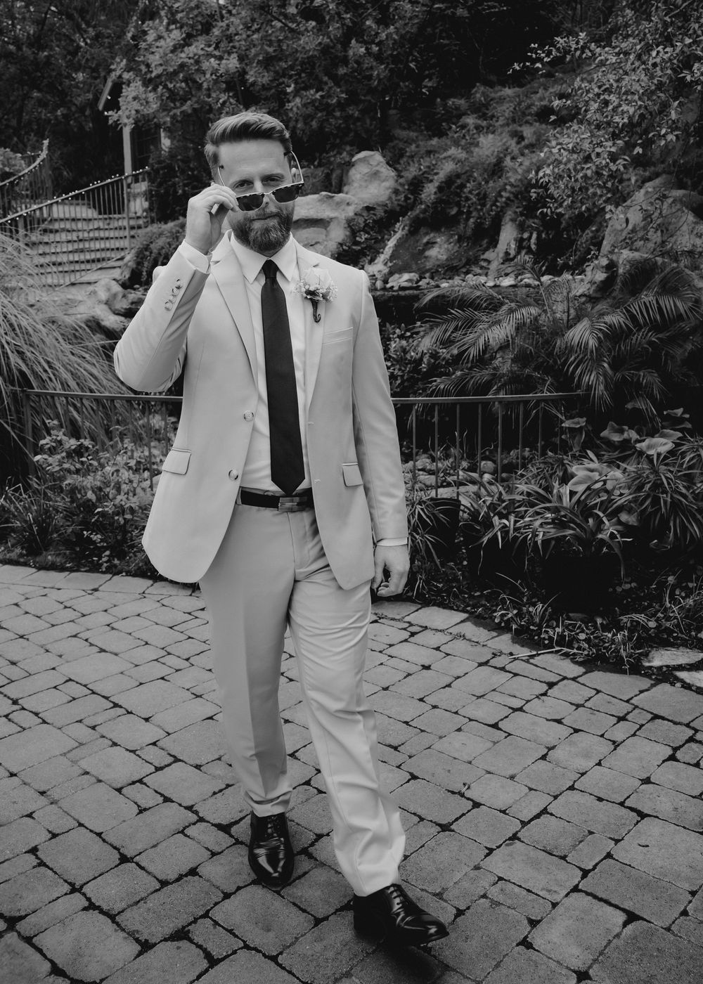 Dapper groom in cream suit and tie adjusts sunglasses while walking on stone patio.