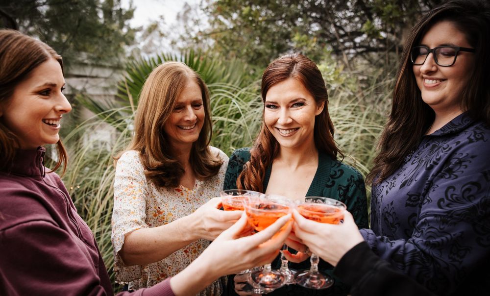 Four women smiling and toasting with pink cocktails at an outdoor celebration.