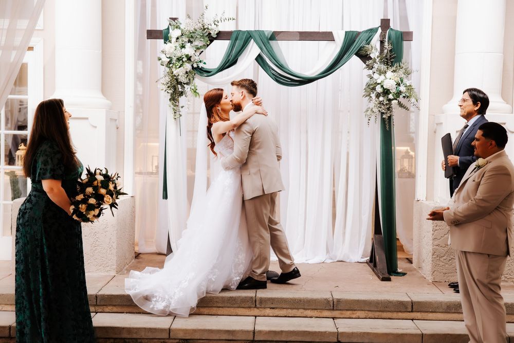 Bride and groom share first kiss beneath a decorated wedding arch with green draping and white flowers.