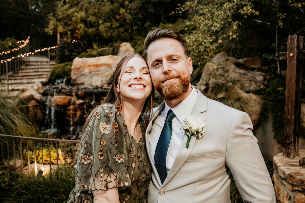 Smiling bride and bearded groom pose together at outdoor wedding venue with waterfall backdrop.