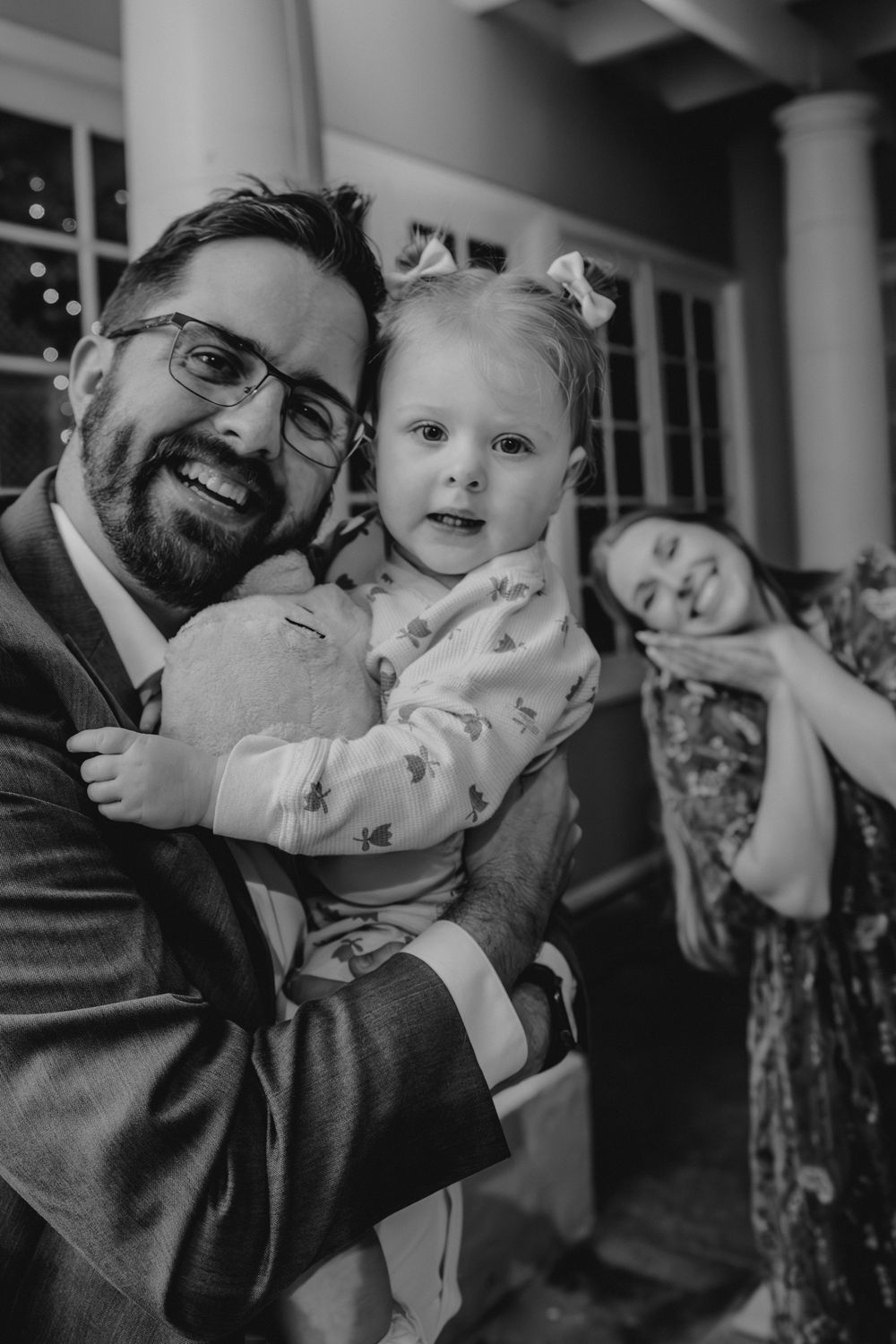 Smiling bearded man in suit holding toddler girl with bow at wedding reception.