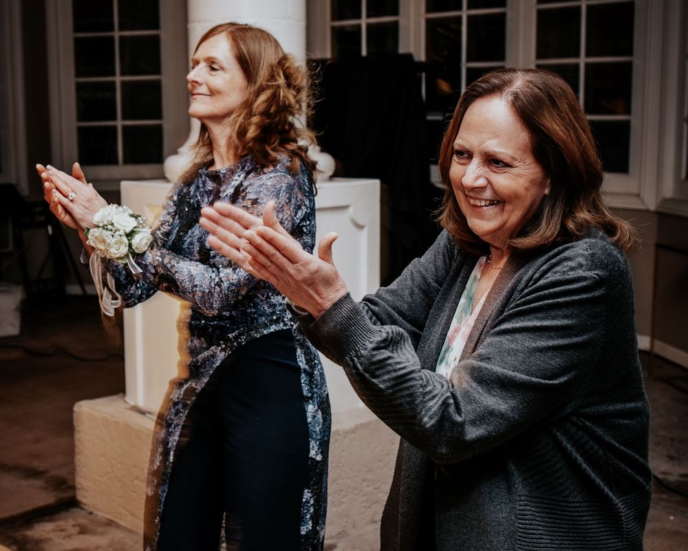 Two women clapping and smiling warmly together at an elegant indoor wedding reception.