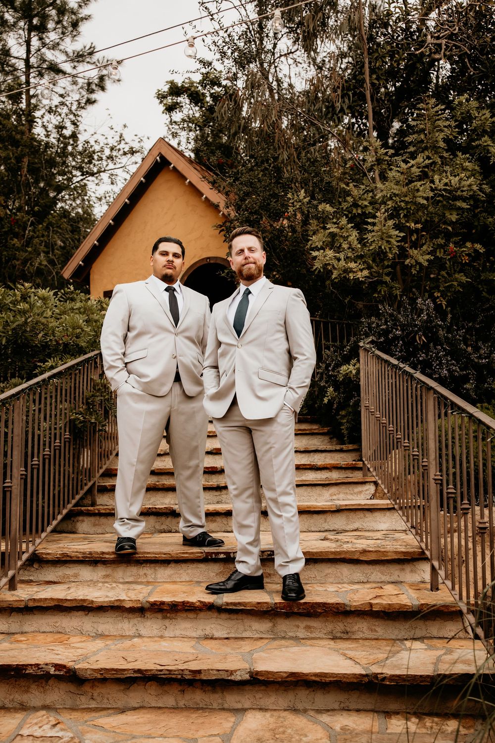 Two grooms in light gray suits with dark ties pose seriously on stone steps before a chapel.