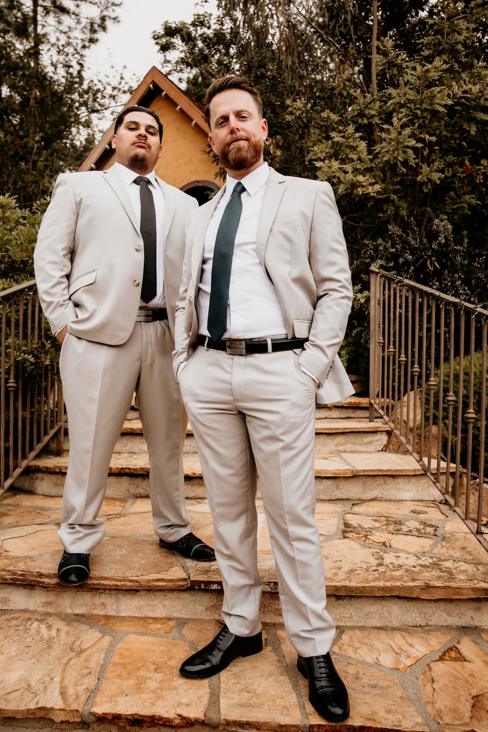 Two well-dressed men in cream suits and dark ties stand confidently on outdoor stone stairs.