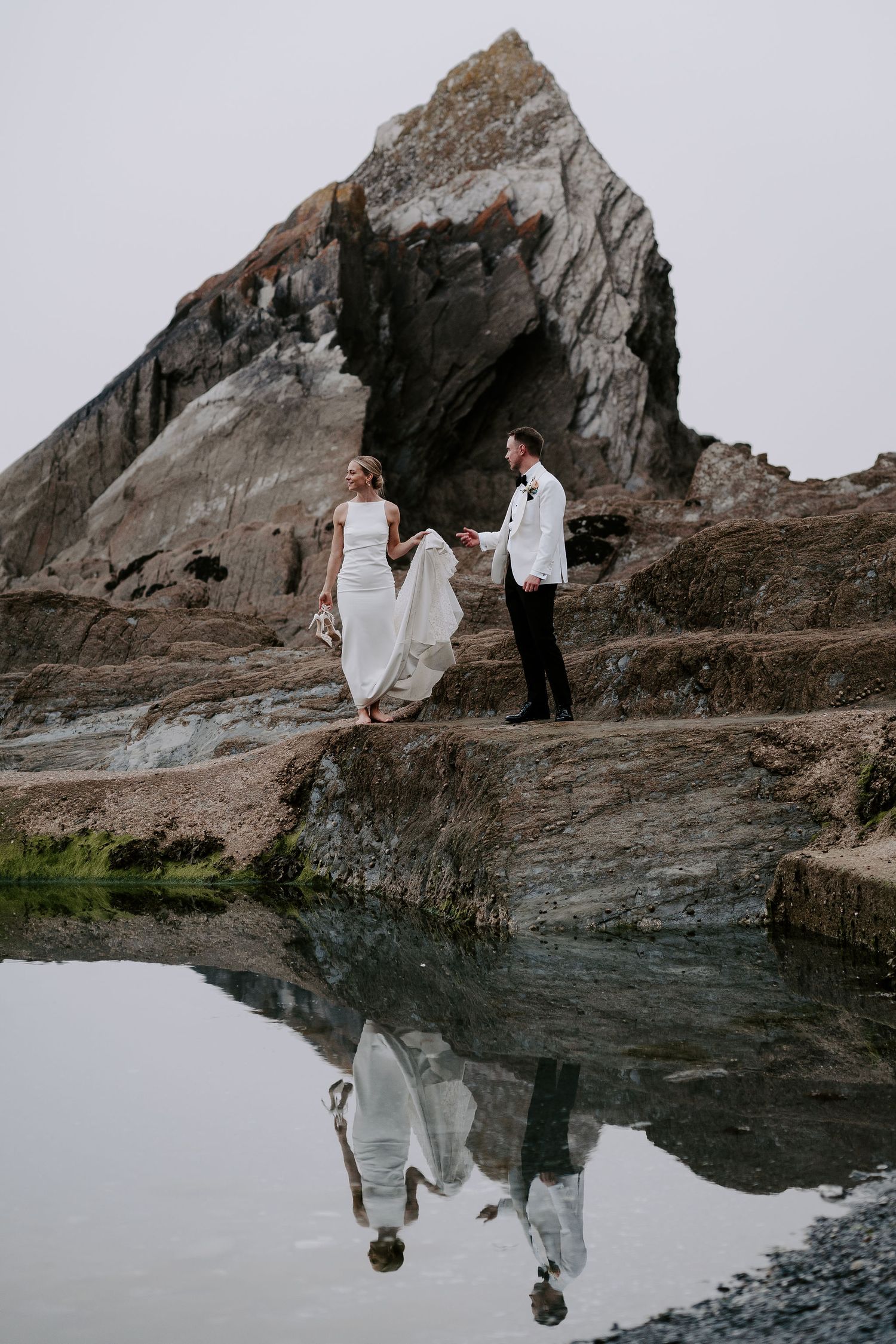 Bride and Groom walking along rocks on beach during sunset at tunnels beach wedding venue in north Devon