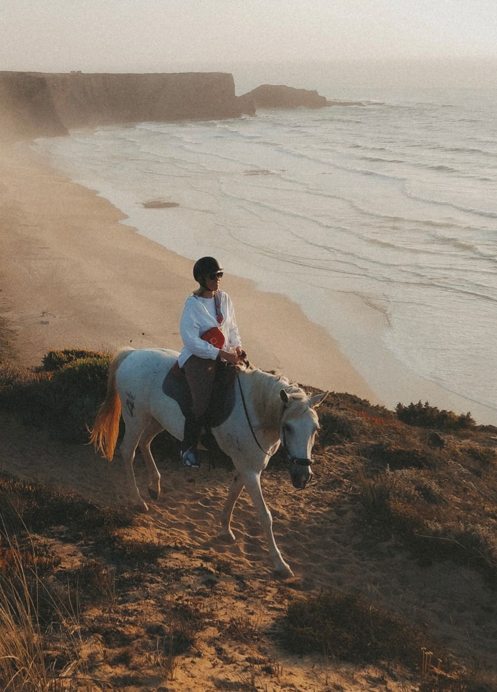 Person riding white horse along dramatic coastal cliffside beach at golden sunset hour.