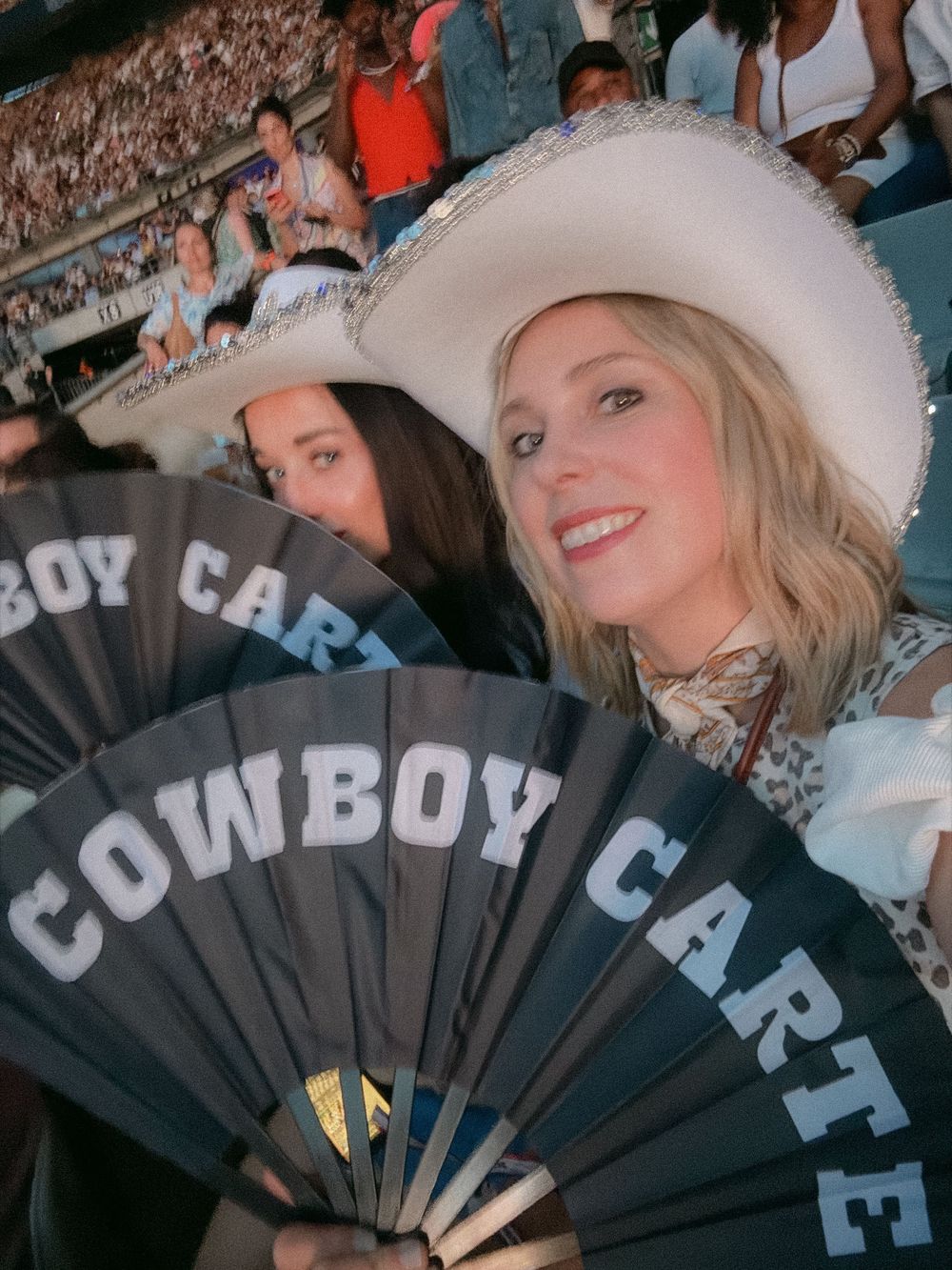 Blonde woman in white cowboy hat holds Cowboy Carter fans at a Beyoncé concert in a packed stadium.